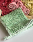 A green hand-dyed hemp exfoliating washcloth with a basket full of rolled hemp exfoliating washcloths in the background. 