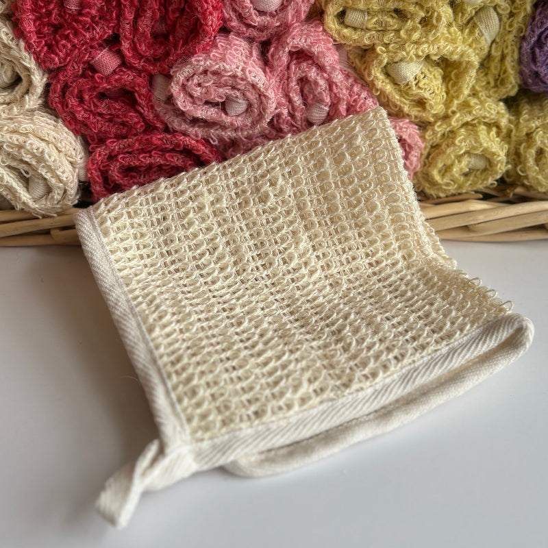 A cream hand-dyed hemp exfoliating washcloth with a basket full of rolled hemp exfoliating washcloths in the background. 
