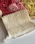 A cream hand-dyed hemp exfoliating washcloth with a basket full of rolled hemp exfoliating washcloths in the background. 