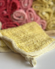 A yellow hand-dyed hemp exfoliating washcloth with a basket full of rolled hemp exfoliating washcloths in the background. 