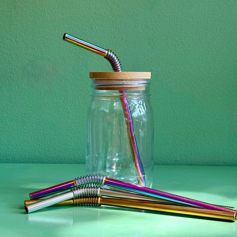 A reused Mason jar with a bamboo straw top and a rainbow bendy metal straw. In front of it sit silver, gold, and rainbow bendy metal straws on a green background.
