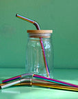 A reused Mason jar with a bamboo straw top and a rainbow bendy metal straw. In front of it sit silver, gold, and rainbow bendy metal straws on a green background.