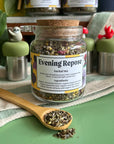 A refillable Jar of Evening repose tea. The jar has a cork lid and an attachable bamboo spoon. The Jar is on a green counter and surrounded by floating tea steepers. 
