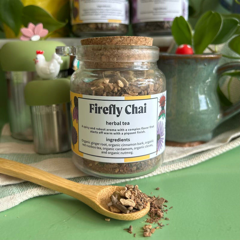 A refillable jar of firefly chai tea. The jar has a cork lid and an attachable bamboo spoon. The spoon is in front of the jar with tea in it. 