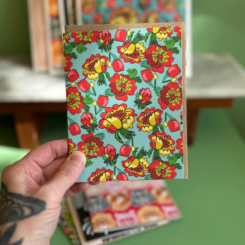 A hand holding a card with a light blue background and bright yellow and red florals. 