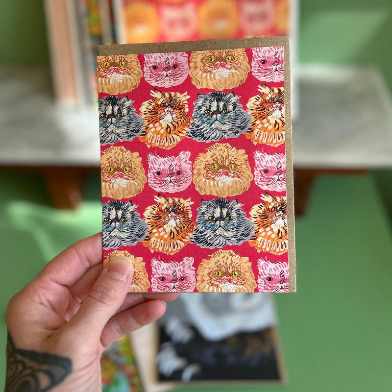 A hand holding a card with a red background and a pattern of several long haired cats.