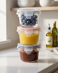 Three borosilicate glass containers in light blue, red, and dark blue stacked on top of each other sitting on a kitchen counter. One holds blueberries, one holds applesauce, and one holds barbecue sauce. 