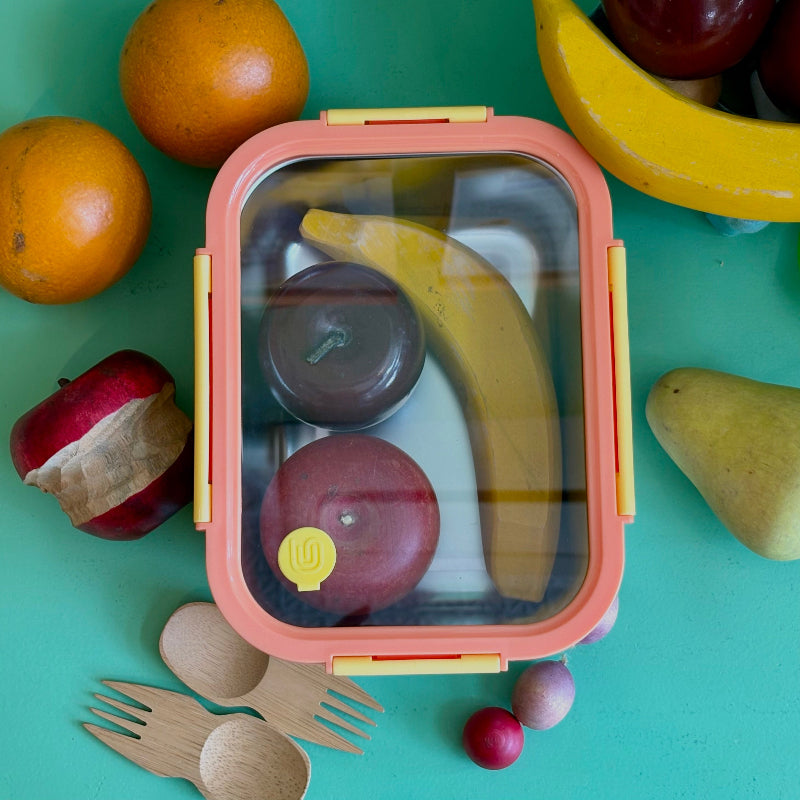 A yellow and orange stainless steel 5 cup food storage container. It is holding wooden apples and bananas and is surrounded by other wooded fruits and bamboo sporks. This all sits on a green table.