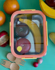 A yellow and orange stainless steel 5 cup food storage container. It is holding wooden apples and bananas and is surrounded by other wooded fruits and bamboo sporks. This all sits on a green table.