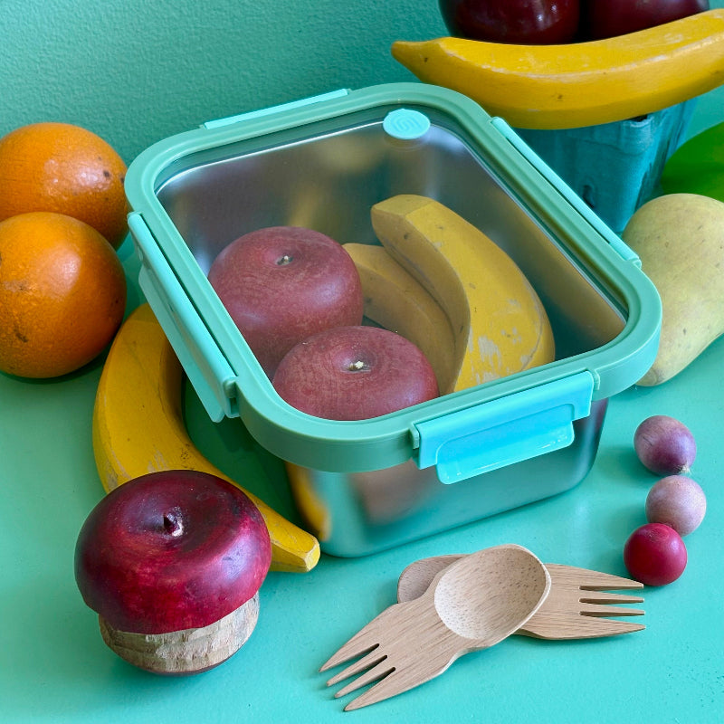 A light blue and green stainless steel 7 and a half cup food storage container. It is holding wooden apples and bananas and is surrounded by other wooded fruits and bamboo sporks. This all sits on a green table.