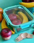 A light blue and green stainless steel 7 and a half cup food storage container. It is holding wooden apples and bananas and is surrounded by other wooded fruits and bamboo sporks. This all sits on a green table.