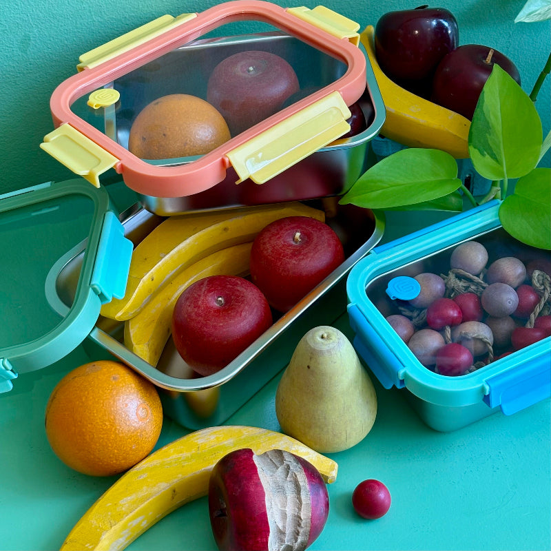 A set of reusable stainless steel containers. Each size has a different colored  glass lid with a steam vent. They are holding wooden fruit on a green background. 