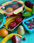 A set of reusable stainless steel containers. Each size has a different colored  glass lid with a steam vent. They are holding wooden fruit on a green background. 