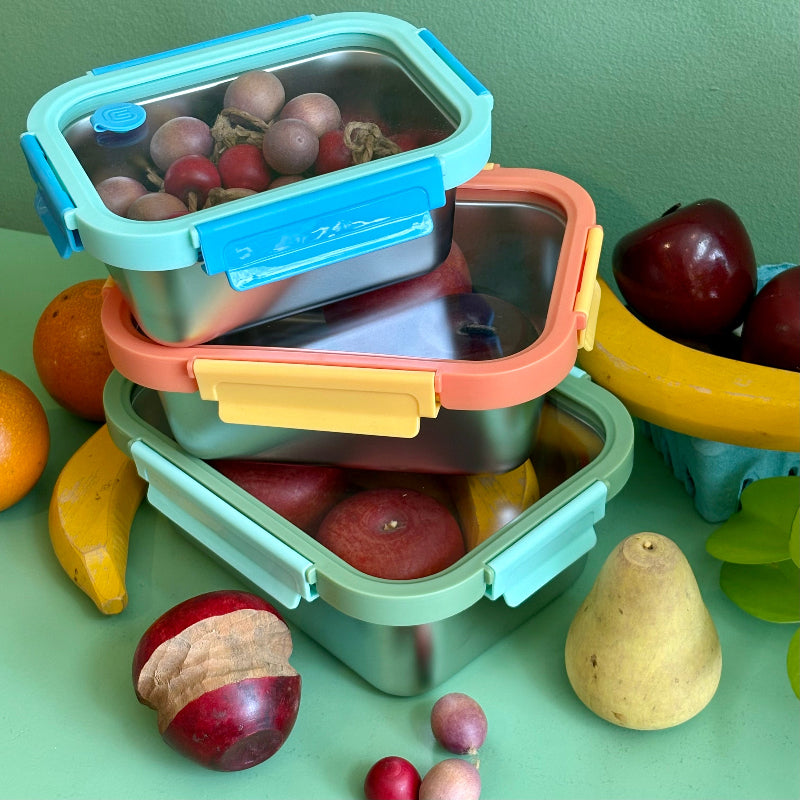 Three stainless steel reusable containers with glass lids stacked on top of each other. They are holding and surrounded by wooden fruits and on a green background.