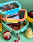 Three stainless steel reusable containers with glass lids stacked on top of each other. They are holding and surrounded by wooden fruits and on a green background.