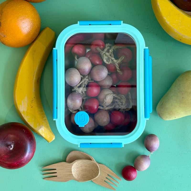 A light blue and dark blue stainless steel 3 and a half cup food storage container. It is holding wooden grapes and is surrounded by other wooded fruits and bamboo sporks. This all sits on a green table.