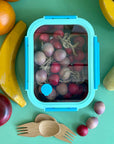 A light blue and dark blue stainless steel 3 and a half cup food storage container. It is holding wooden grapes and is surrounded by other wooded fruits and bamboo sporks. This all sits on a green table.