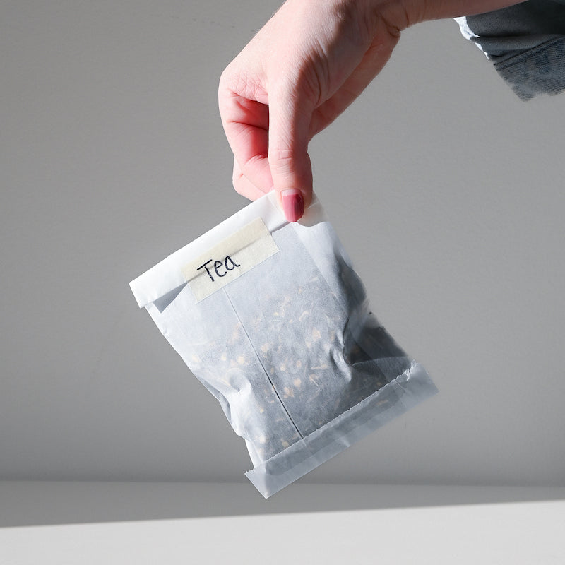 A hand holds a white paper bag labeled  tea against a plain light gray background.