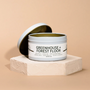 A white Modern Makers Modern Maker Provisions Soy Candle tin, labeled Greenhouse + Forest Floor, sits with its lid behind it on a textured beige platform against a beige background. This hand-poured soy candle delivers long-lasting, nature-inspired fragrance.