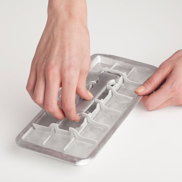 A person's hands are shown pressing down on a silver metal ice cube tray mold to demonstrate its use, set against a clean, light off-white background.