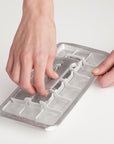 A person's hands are shown pressing down on a silver metal ice cube tray mold to demonstrate its use, set against a clean, light off-white background.