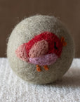 A grey felted wool dryer ball featuring red small bird design, resting on a textured beige fabric surface against a brown background.