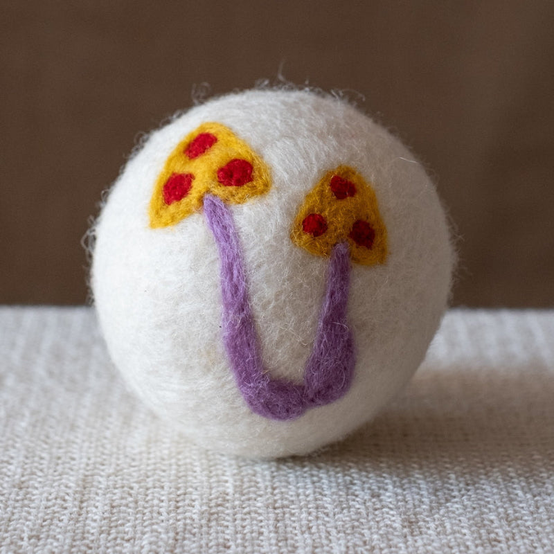 A white wool dryer ball with a handmade  design featuring two yellow mushrooms with red spots and purple stems, resting on a textured beige fabric surface against a solid brown background.