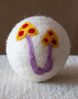A white wool dryer ball with a handmade  design featuring two yellow mushrooms with red spots and purple stems, resting on a textured beige fabric surface against a solid brown background.