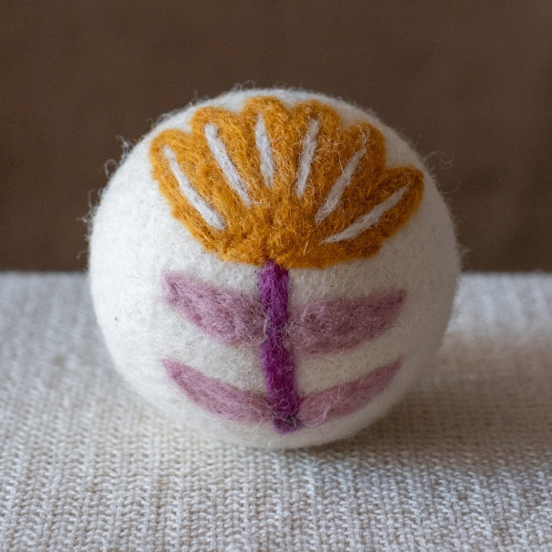 A white felted wool dryer ball featuring a mustard yellow folk art flower with a purple stem, and purple leaves, resting on a textured beige fabric surface against a brown background.
