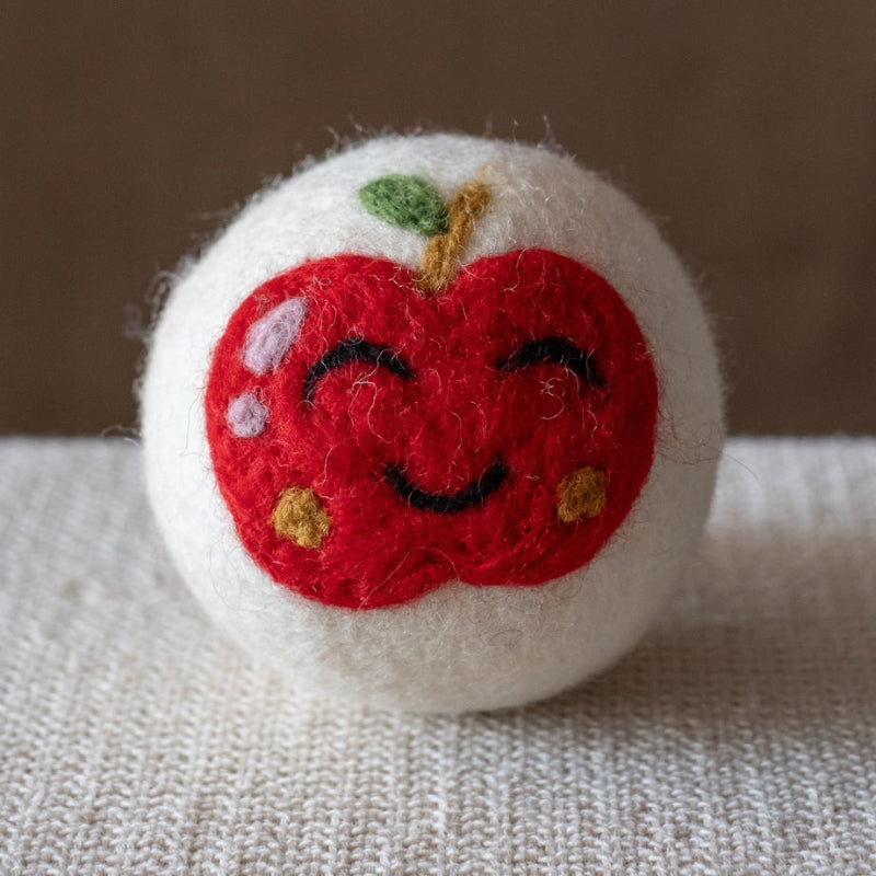 A white felted wool dryer ball featuring a happy red apple design, resting on a textured beige fabric surface against a brown background.