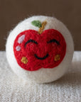 A white felted wool dryer ball featuring a happy red apple design, resting on a textured beige fabric surface against a brown background.