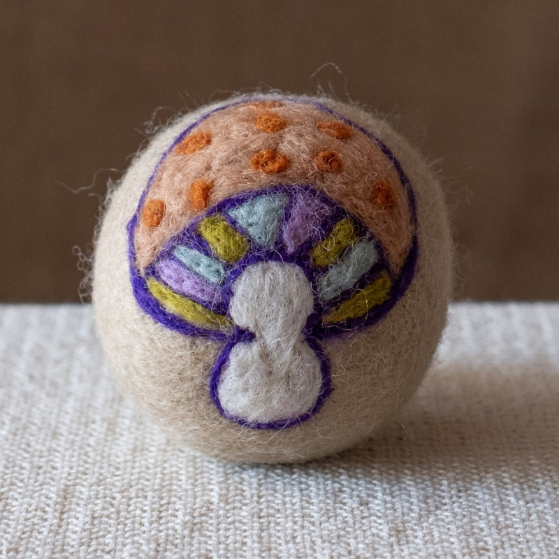 A beige felted wool dryer ball featuring a 70's inspired mushroom design with an orange polka dotted cap, green blue and purple gills, and a white stem. The dryer ball is resting on a textured beige fabric surface against a brown background.