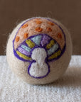 A beige felted wool dryer ball featuring a 70's inspired mushroom design with an orange polka dotted cap, green blue and purple gills, and a white stem. The dryer ball is resting on a textured beige fabric surface against a brown background.