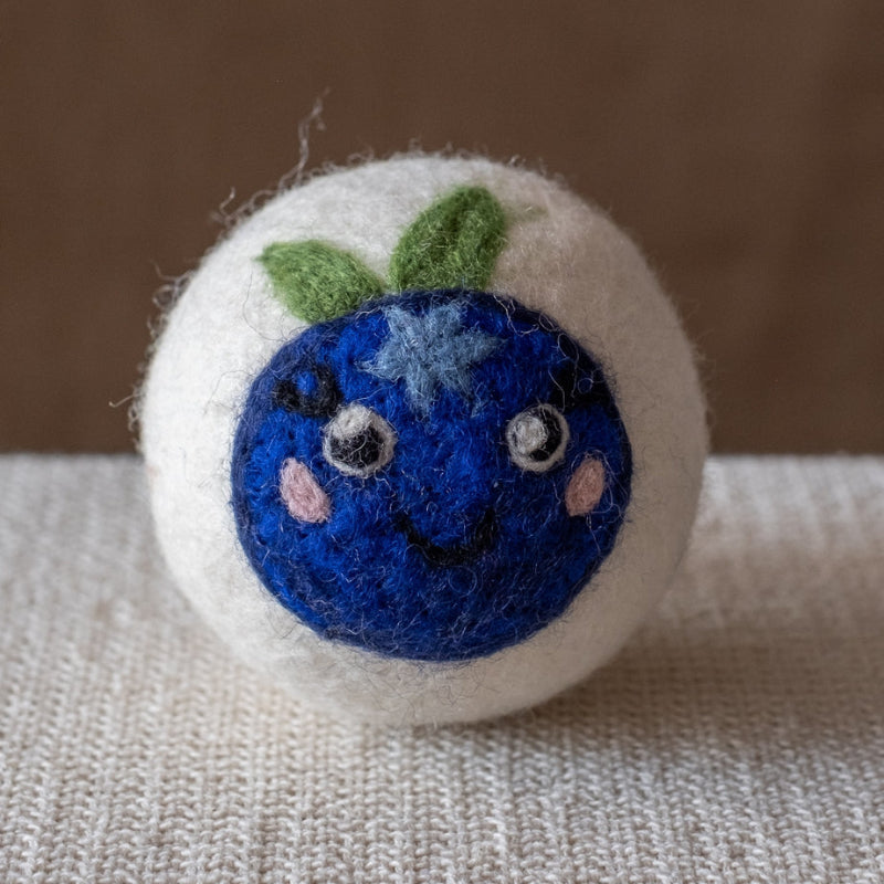 A white felted wool dryer ball featuring a happy blueberry design, resting on a textured beige fabric surface against a brown background.