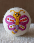 A white felted wool dryer ball featuring a happy purple and yellow butterfly design, resting on a textured beige fabric surface against a brown background.