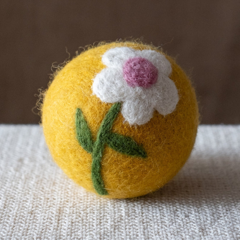 A yellow felted wool dryer ball featuring white daisy, with a green stem, and a pink center, resting on a textured beige fabric surface against a brown background.