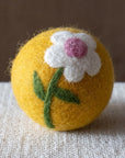 A yellow felted wool dryer ball featuring white daisy, with a green stem, and a pink center, resting on a textured beige fabric surface against a brown background.