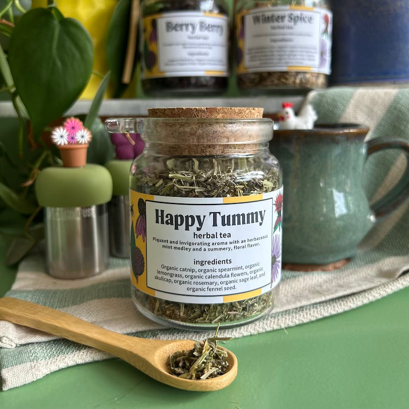 A refillable jar of happy tummy tea. The jar has a cork lid and an attachable bamboo spoon. The spoon is in front of the jar with tea in it. 