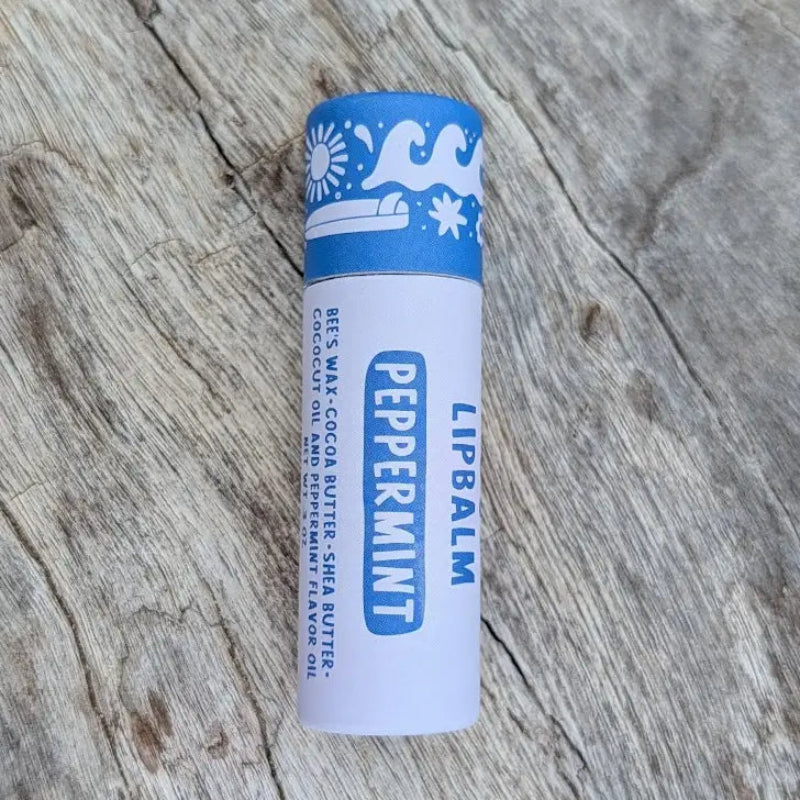 A Peppermint Hawaiian Lip balm in a sustainable white paper tube with blue designs on it, sitting on a wooden surface.