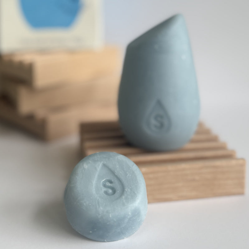 The blue mini hibar moisturize shampoo leaning on a soap dish in front of the full size shampoo bar.