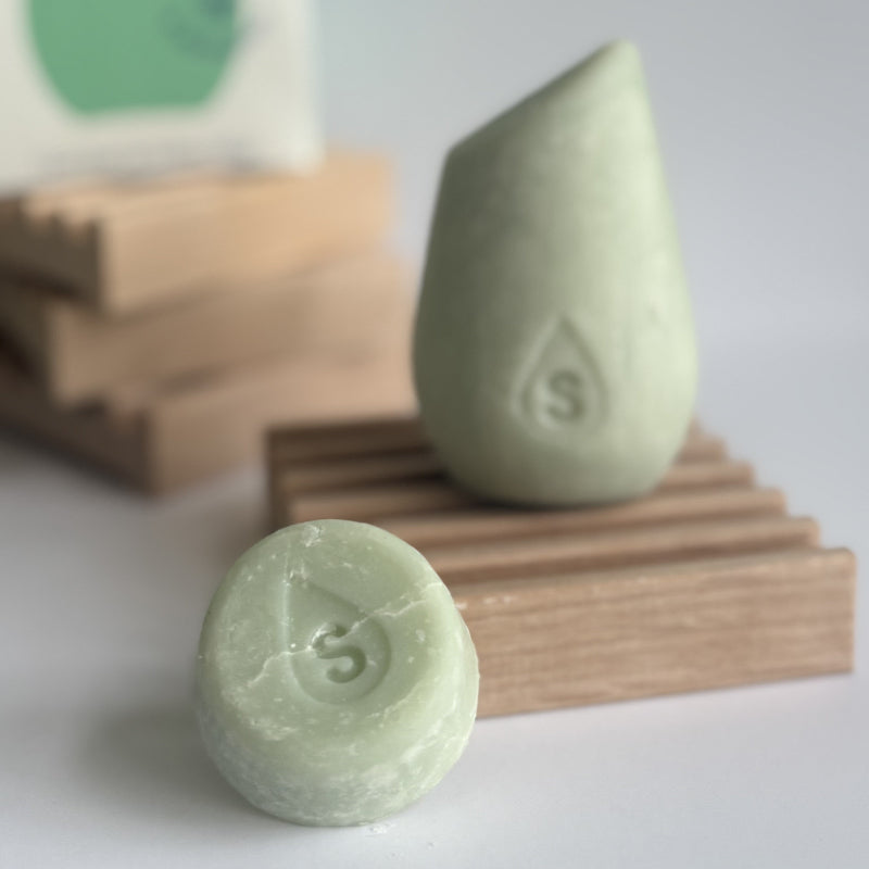 The green mini hibar maintain shampoo leaning on a soap dish in front of the full size shampoo bar.
