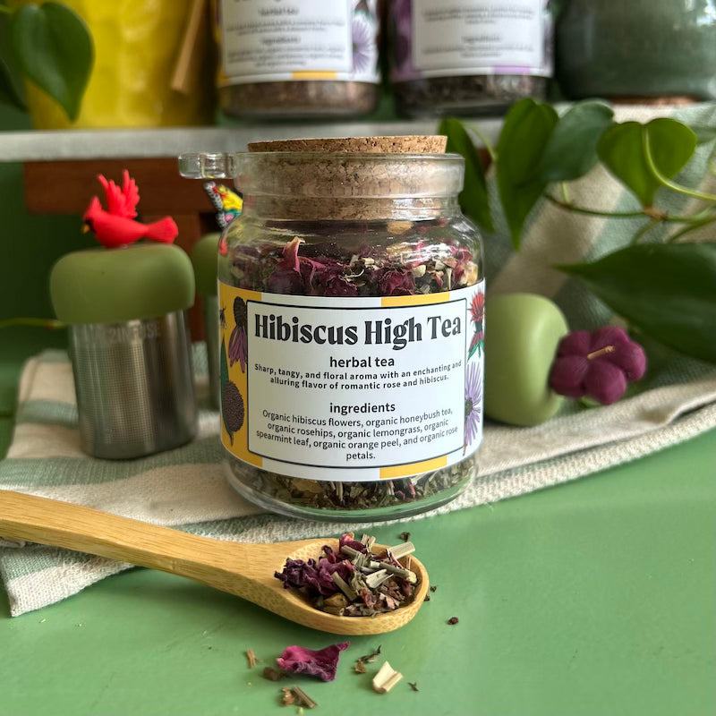 A refillable jar of Hibiscus high tea. The jar has a cork lid and an attachable bamboo spoon. The spoon is in front with tea in it. 