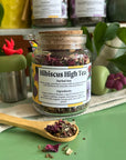 A refillable jar of Hibiscus high tea. The jar has a cork lid and an attachable bamboo spoon. The spoon is in front with tea in it. 