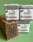 Three wrapped bars of kitchen and gardeners soap by Jangle Soap Works.  The bars are on a green background with a bulk loaf of soap showing the exfoliating details in the soap. 