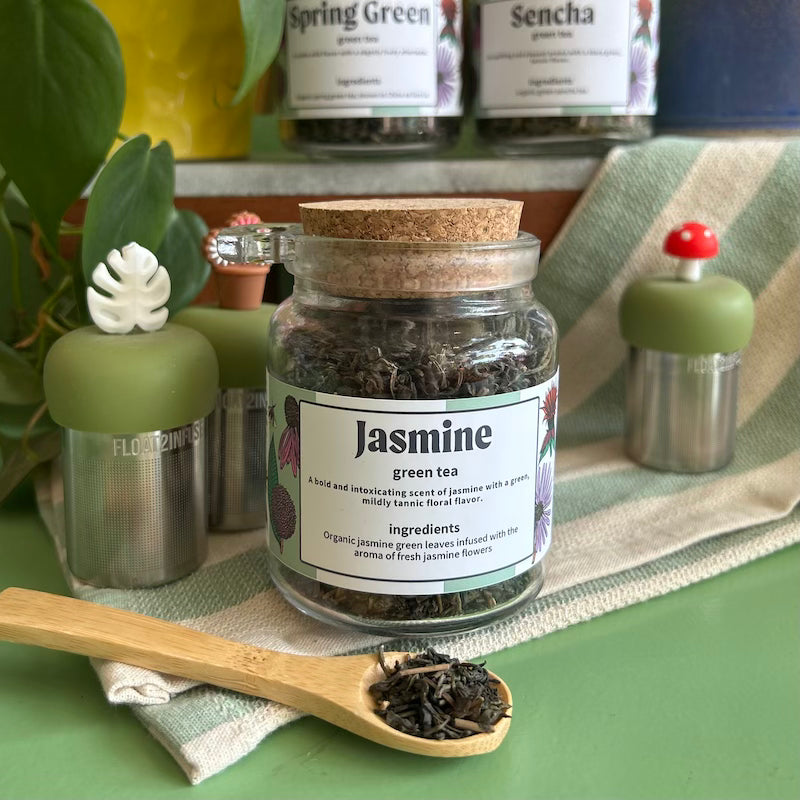 A refillable jar of Jasmine tea. The jar has a cork lid and a bamboo attachable spoon. The spoon is in front of the jar full of jasmine tea. 
