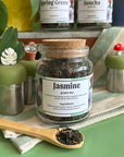 A refillable jar of Jasmine tea. The jar has a cork lid and a bamboo attachable spoon. The spoon is in front of the jar full of jasmine tea. 