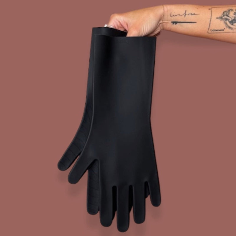 A tattoed hand holds black silicone kitchen gloves on a brown background. 