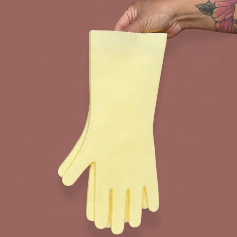 A tattooed hand holds butter yellow silicone kitchen gloves on a brown background. 