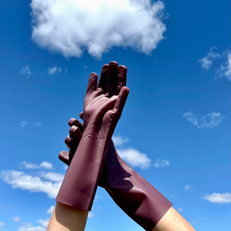 Hands modeling fig colored silicone kitchen gloves. The sturdy, washable kitchen gloves have grooves on the palm and fingers to make handling wet dishes easy. The hands wearing the gloves are lifted into a blue sky. 