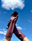 Hands modeling fig colored silicone kitchen gloves. The sturdy, washable kitchen gloves have grooves on the palm and fingers to make handling wet dishes easy. The hands wearing the gloves are lifted into a blue sky. 